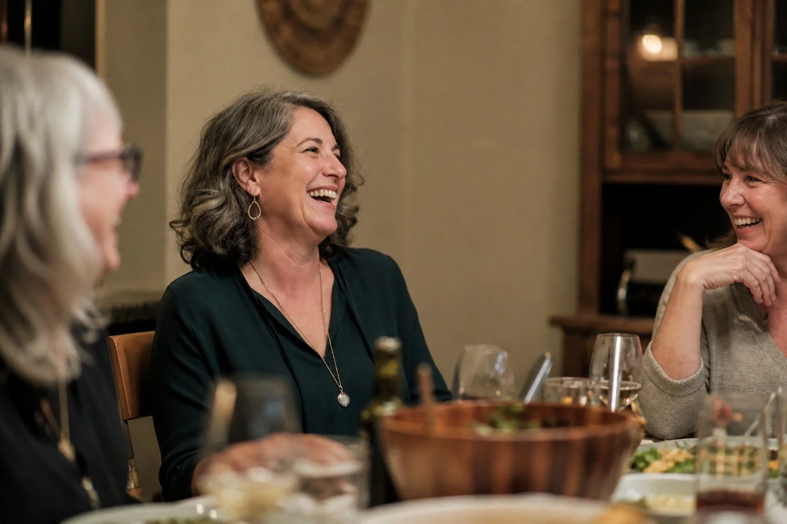 A woman in her fifties laughing naturally with close friends over dinner.